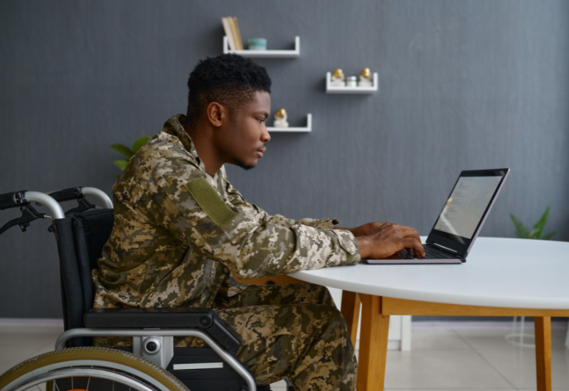 Man in military uniform sitting in a wheelchair, at a table using a laptop